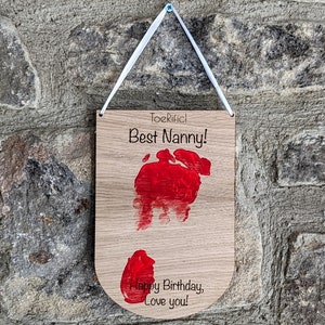 K&ouml;nnte beinhalten: Ein Holzschild mit einem roten Handabdruck und dem Text "Toerific! Best Nanny! Happy Birthday, Love you!" h&auml;ngt an einer Steinwand.