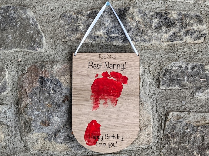 K&ouml;nnte beinhalten: Ein Holzschild mit einem roten Handabdruck und dem Text "Toerific! Best Nanny! Happy Birthday, Love you!" h&auml;ngt an einer Steinwand.
