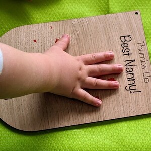 K&ouml;nnte beinhalten: Eine Holzplakette mit dem Text "Thumbs Up Best Nanny!" Ein roter Handabdruck eines Kindes befindet sich auf der Plakette.