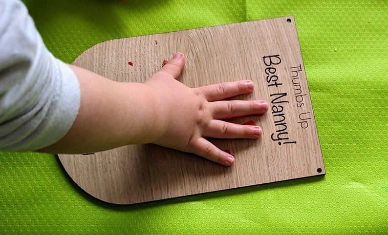 K&ouml;nnte beinhalten: Eine Holzplakette mit dem Text "Thumbs Up Best Nanny!" Ein roter Handabdruck eines Kindes befindet sich auf der Plakette.