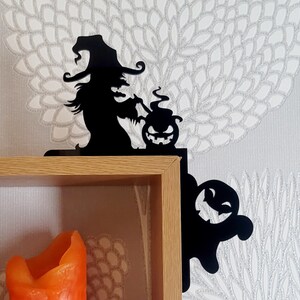 May include: Black metal shelf brackets with a witch and a ghost design. The witch is holding a cauldron with a jack-o-lantern inside. The ghost is smiling. The brackets are mounted on a wooden shelf with a candle inside.