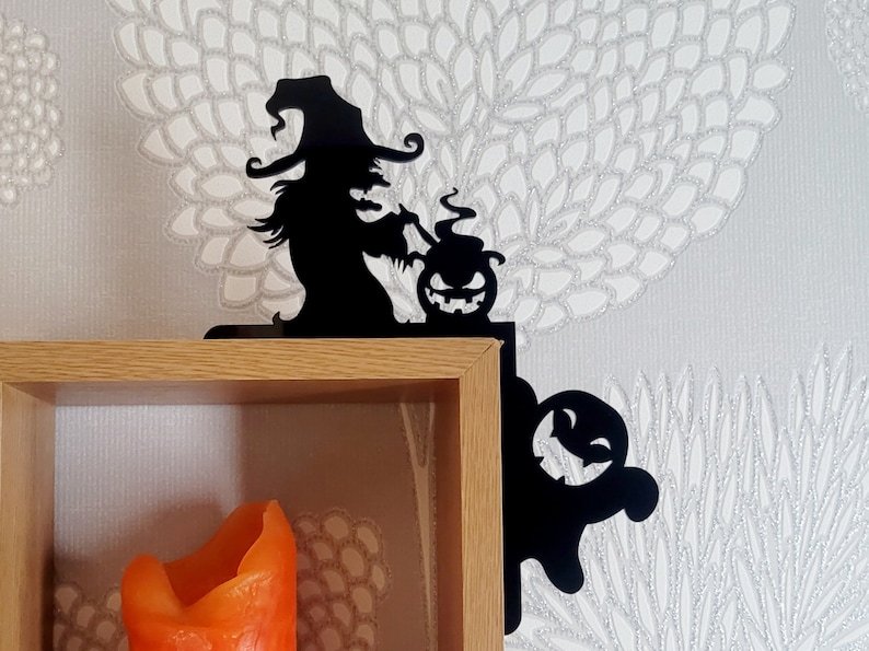 May include: Black metal shelf brackets with a witch and a ghost design. The witch is holding a cauldron with a jack-o-lantern inside. The ghost is smiling. The brackets are mounted on a wooden shelf with a candle inside.