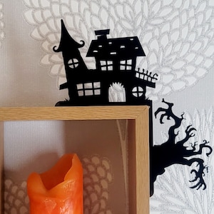 May include: Black metal Halloween shelf bracket shaped like a haunted house with a tree branch. The bracket is mounted on a wooden shelf with an orange candle inside.