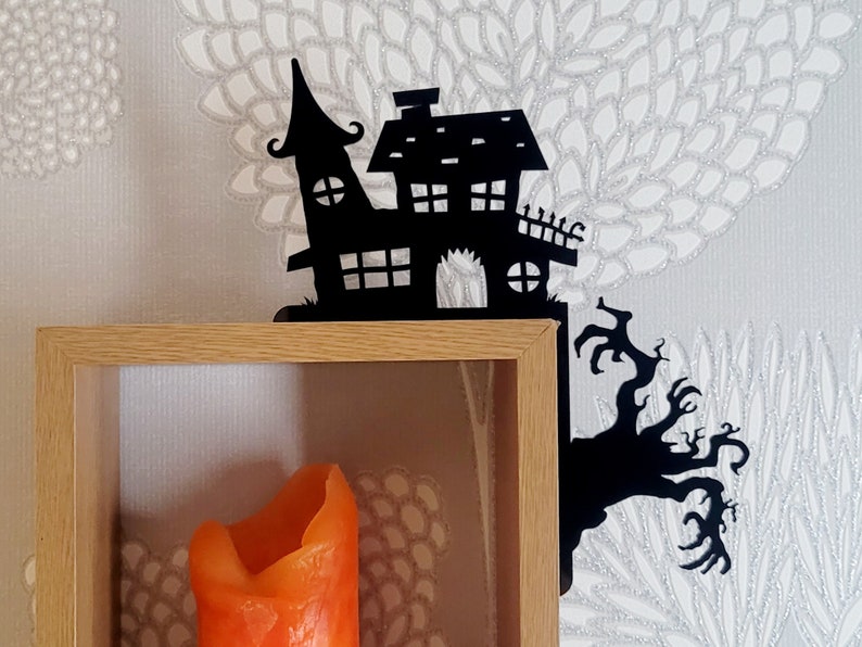 May include: Black metal Halloween shelf bracket shaped like a haunted house with a tree branch. The bracket is mounted on a wooden shelf with an orange candle inside.