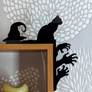 May include: Black silhouette of a cat, a witch's hat, and a hand reaching out from a tree, mounted on a wooden shelf.