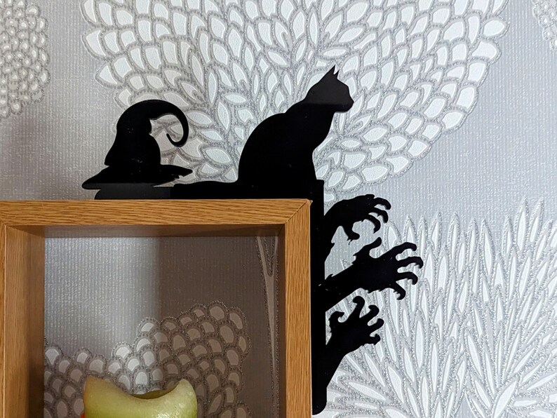 May include: Black silhouette of a cat, a witch's hat, and a hand reaching out from a tree, mounted on a wooden shelf.