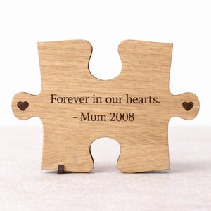 Puede incluir: Una pieza de rompecabezas de madera de color marrón claro con el texto grabado "Forever in our hearts. - Mum 2008" centrado en el frente. Pequeños iconos de corazón de color marrón oscuro están grabados a los lados.