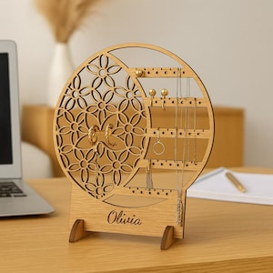 Puede incluir: Un organizador de joyas de madera con un diseño floral y el nombre "Olivia" grabado en la base. El organizador tiene múltiples agujeros para pendientes y ganchos para collares. La madera es de color marrón claro.