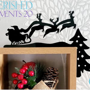 May include: Black metal silhouette of Santa Claus in a sleigh pulled by reindeer flying over a Christmas tree. The silhouette is mounted on a wooden shelf with a Christmas-themed decoration in the foreground.