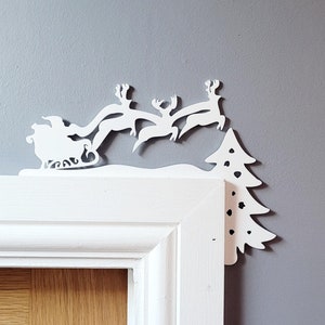 May include: White paper cutout of Santa Claus in a sleigh pulled by reindeer flying over a white paper cutout of a Christmas tree. The cutout is attached to a white door frame.