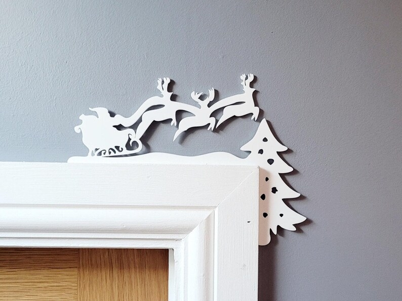 May include: White paper cutout of Santa Claus in a sleigh pulled by reindeer flying over a white paper cutout of a Christmas tree. The cutout is attached to a white door frame.