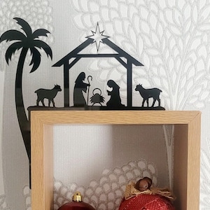 May include: Black metal silhouette of a nativity scene with a palm tree, two sheep, Mary, Joseph, and baby Jesus in a manger. The silhouette is mounted on a wooden shelf.
