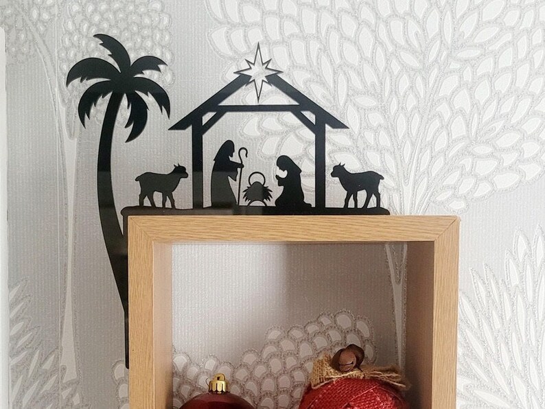 May include: Black metal silhouette of a nativity scene with a palm tree, two sheep, Mary, Joseph, and baby Jesus in a manger. The silhouette is mounted on a wooden shelf.