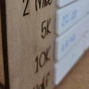 Może przedstawiać: Drewniany tracker odległości do biegania z wytrawionym tekstem. Odległości obejmują 2 Mile, 5K, 10K i Half. Białe bloki z niebieskim tekstem po prawej stronie. Drewniana tablica jest jasnobrązowa.