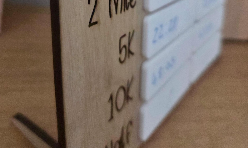 Może przedstawiać: Drewniany tracker odległości do biegania z wytrawionym tekstem. Odległości obejmują 2 Mile, 5K, 10K i Half. Białe bloki z niebieskim tekstem po prawej stronie. Drewniana tablica jest jasnobrązowa.