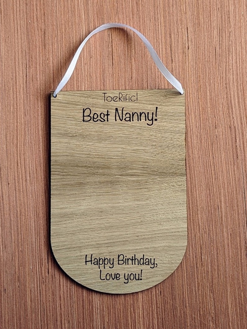K&ouml;nnte beinhalten: Holzschilder mit einem wei&szlig;en Band zum Aufh&auml;ngen. Das Schild lautet "Toerific! Best Nanny! Happy Birthday, Love you!"