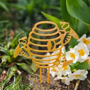 May include: A gold metal beehive shaped garden stake with two bees on either side. The stake is in a garden setting with white flowers.