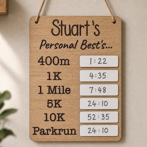 Può includere: Cartello in legno chiaro con la scritta "Stuart's Personal Best's..." e i tempi di corsa per 400m, 1K, 1 Miglio, 5K, 10K e Parkrun. Il cartello è appeso con una corda. Le misure sono in metri e chilometri.