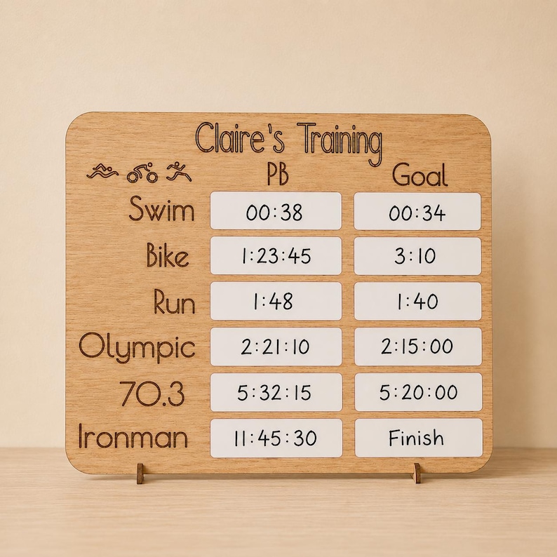 Personalised triathlon training tracker board in oak veneer wood with engraved “Claire’s Training” heading, white dry wipe PB and goal sections, and handwritten race times and Ironman goals. The handmade wooden board is displayed on thin wooden stands against a soft beige wall with warm natural lighting and a clean minimalist Etsy-style setup.