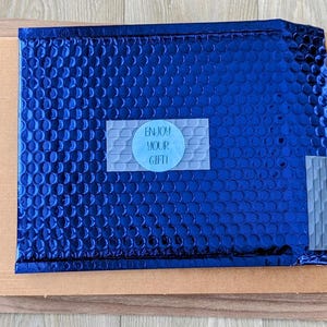 Może przedstawiać: Niebieska koperta bąbelkowa z naklejką z napisem "Enjoy Your Gift!" umieszczona w kartonowym pudełku. Koperta ma teksturowaną powierzchnię i jest zaklejona taśmą. Pudełko znajduje się na drewnianej powierzchni.
