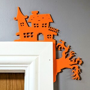 May include: Orange Halloween-themed house and tree cutout decoration. The house has a pointed roof and windows. The tree has branches with spooky, claw-like fingers.
