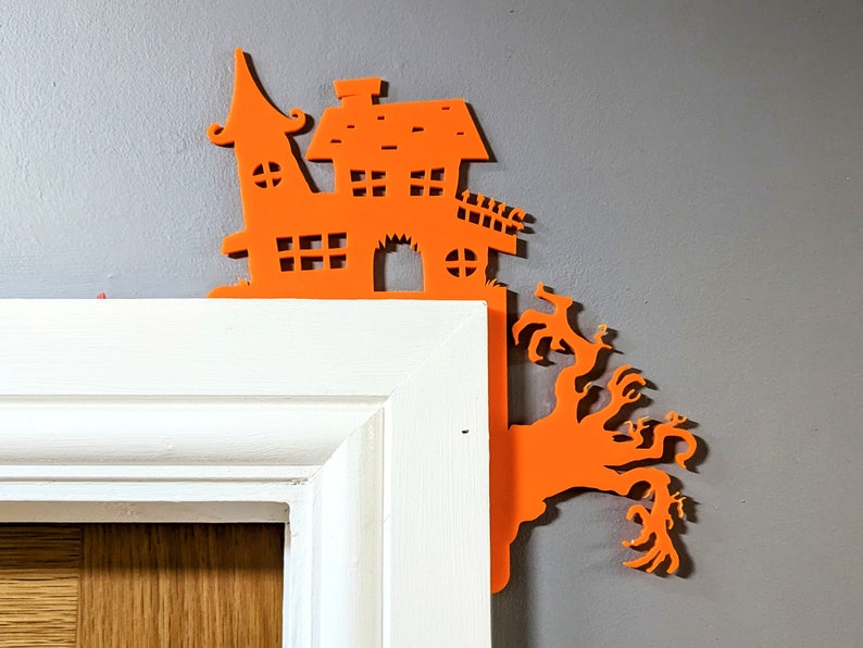 May include: Orange Halloween-themed house and tree cutout decoration. The house has a pointed roof and windows. The tree has branches with spooky, claw-like fingers.