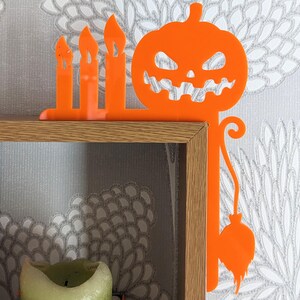 May include: Orange Halloween-themed shelf decor with a pumpkin and candles. The pumpkin has a scary face and a stem with a dangling ball. The candles are in a row and are tall and thin.