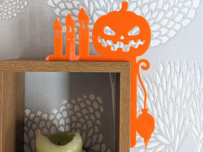 May include: Orange Halloween-themed shelf decor with a pumpkin and candles. The pumpkin has a scary face and a stem with a dangling ball. The candles are in a row and are tall and thin.