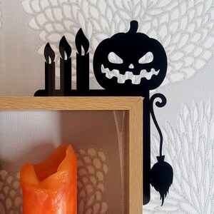 May include: Black metal shelf bracket with a pumpkin and three candles design. The bracket is attached to a wooden shelf with a lit orange candle inside.