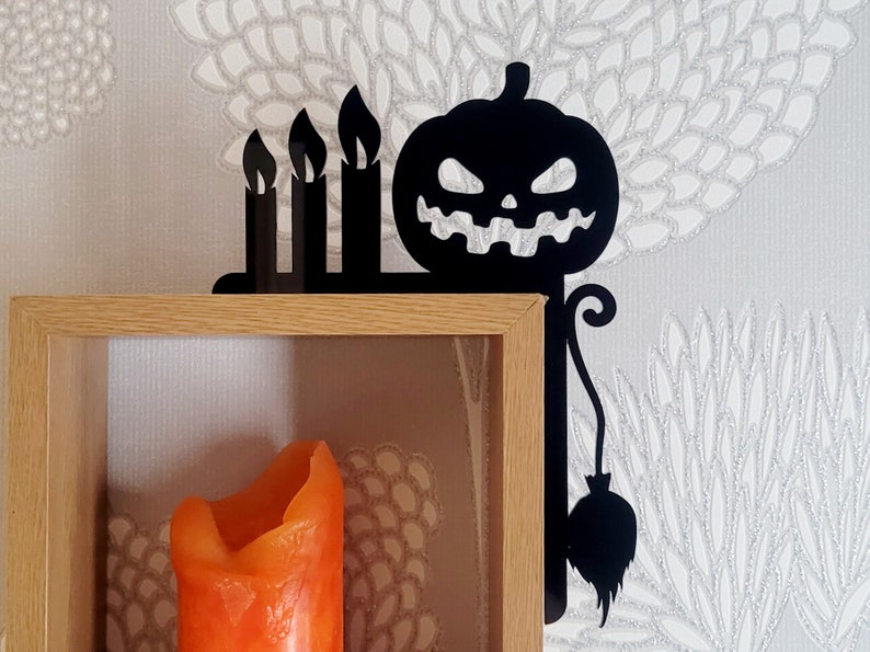 May include: Black metal shelf bracket with a pumpkin and three candles design. The bracket is attached to a wooden shelf with a lit orange candle inside.