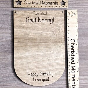 K&ouml;nnte beinhalten: Holzzeichen mit dem Text "ToeRificl Best Nanny!" und "Happy Birthday, Love you!" Das Schild ist an einem Holzlineal mit dem Text "Cherished Moments" befestigt.