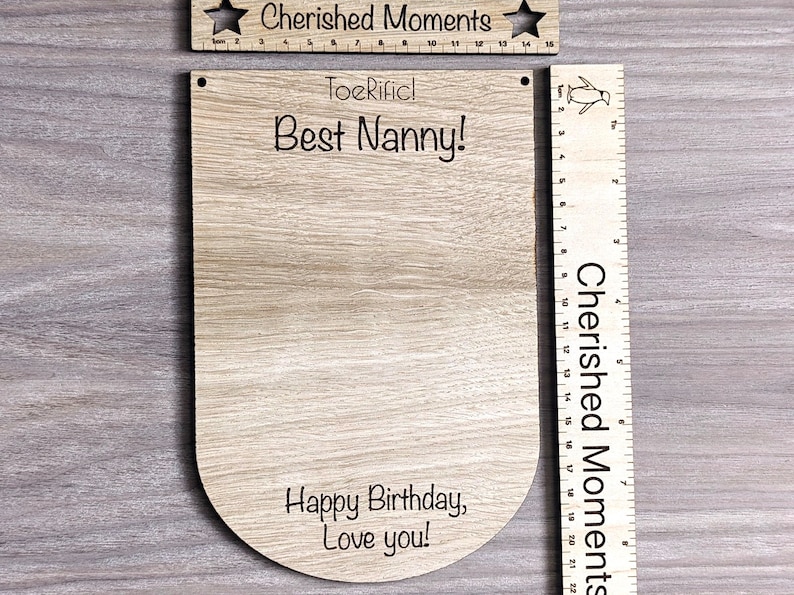 K&ouml;nnte beinhalten: Holzzeichen mit dem Text "ToeRificl Best Nanny!" und "Happy Birthday, Love you!" Das Schild ist an einem Holzlineal mit dem Text "Cherished Moments" befestigt.