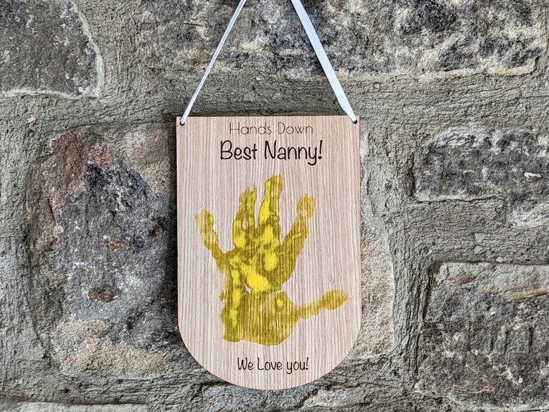 K&ouml;nnte beinhalten: Holzschilder mit dem Text "Hands Down Best Nanny!" und einem gelben Handabdruck. Das Schild sagt auch "We Love you!"