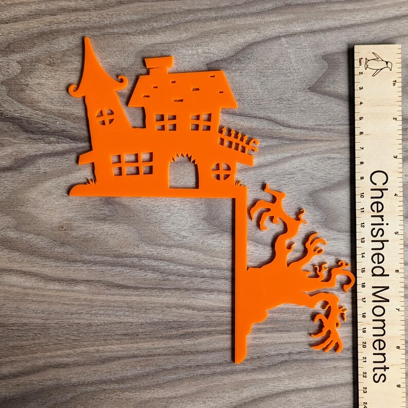 May include: Orange Halloween-themed cutout of a haunted house with a spooky tree. The house has a pointed roof, windows, and a door. The tree has gnarled branches and is partially obscured by the house.