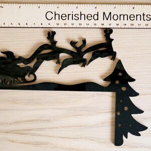 May include: Black silhouette of Santa Claus in a sleigh pulled by reindeer flying over a black silhouette of a Christmas tree. The image has the text "Cherished Moments" above the silhouette.