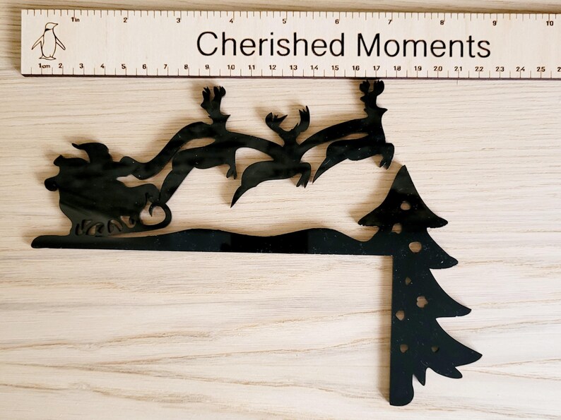 May include: Black silhouette of Santa Claus in a sleigh pulled by reindeer flying over a black silhouette of a Christmas tree. The image has the text "Cherished Moments" above the silhouette.