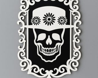 Op de afbeelding: Zwart-wit wanddecoratie met een schedel die een hoed met bloemen draagt. De schedel staat tegen een zwarte achtergrond in een witte, sierlijke lijst. Het ontwerp is een unieke mix van gotische en bloemelementen.