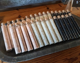 May include: A tray of small glass vials filled with different types of salt. The vials are arranged in rows and are labeled with cork stoppers. The salt is white, pink, and black.