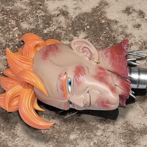 May include: A sculpted figurine head of a character with spiky orange hair, light skin, and blue eyes. The face features red painted battle damage marks, a small gold hoop earring, and a metallic silver neck connector with protruding metal pins.