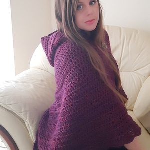 Puede incluir: Un poncho de ganchillo morado con capucha y un patrón de punto texturizado. El poncho está drapeado sobre un sofá blanco.