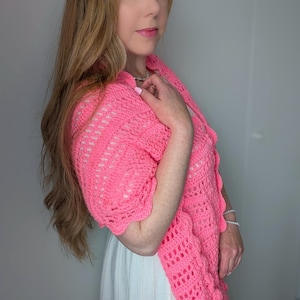 May include: A vibrant pink, open-weave crochet shawl with a scalloped edge. The shawl is draped over the shoulders, highlighting the intricate stitch pattern and colour. A handmade accessory.