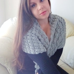 May include: A woman wearing a grey crocheted shawl with a black long-sleeved shirt. She is sitting on a white sofa.