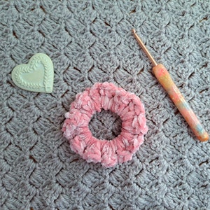 May include: A pink fuzzy hair scrunchie, a white heart-shaped decorative item, and a pink and gold crochet hook on a gray crocheted surface.