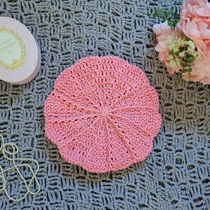 May include: A handmade, light pink crochet beret hat. The hat is circular with a scalloped edge. A pink and white striped oval box with gold text is in the background, along with a string of pearls and pink flowers.