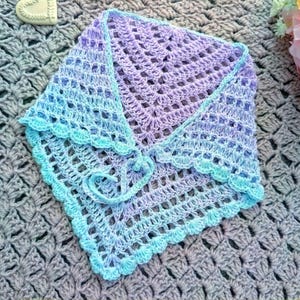 May include: A handmade, crocheted shawl in shades of lavender, blue, and turquoise. The shawl features a delicate openwork design with a scalloped edge and a tie closure. The shawl is approximately 30 inches wide.