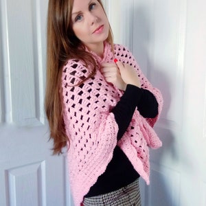 May include: A pink crocheted shawl with a lacy pattern. The shawl is draped over the shoulders of a person wearing a black mock neck shirt and a plaid skirt.