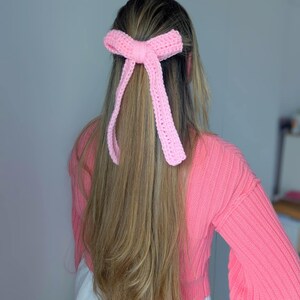 Peut inclure: Un accessoire pour cheveux en forme de n&oelig;ud rose clair, crochet&eacute;. Le n&oelig;ud est attach&eacute; dans les cheveux, avec de longs rubans pendants. L'accessoire est fait d'un fil doux et textur&eacute;. La personne porte un pull rose.