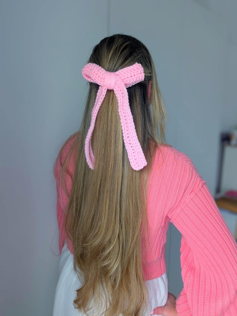 Peut inclure: Un accessoire pour cheveux en forme de n&oelig;ud rose clair, crochet&eacute;. Le n&oelig;ud est attach&eacute; dans les cheveux, avec de longs rubans pendants. L'accessoire est fait d'un fil doux et textur&eacute;. La personne porte un pull rose.