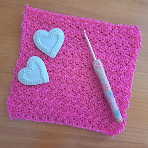 Puede incluir: Un cuadrado de crochet rosa con un gancho de crochet blanco y dos adornos en forma de corazón blanco.