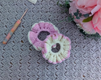 May include: Two handmade crochet scrunchies in pink and cream with a scalloped edge, next to a crochet hook with a colorful handle. The scrunchies are on a gray crocheted surface, with pink flowers in the background.